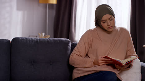Muslim-Woman-Wearing-Hijab-Sitting-On-Sofa-At-Home-Reading-Or-Studying-The-Quran-1